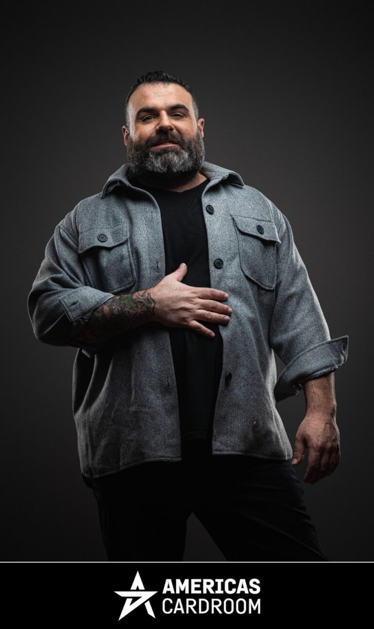 A medium shot of Jon Van Fleet, an ACR poker pro, with a full beard, looking directly at the camera with a serious expression. He is wearing a grey button-up overshirt over a black t-shirt, black pants, and has his left hand resting on his chest, against a dark background.