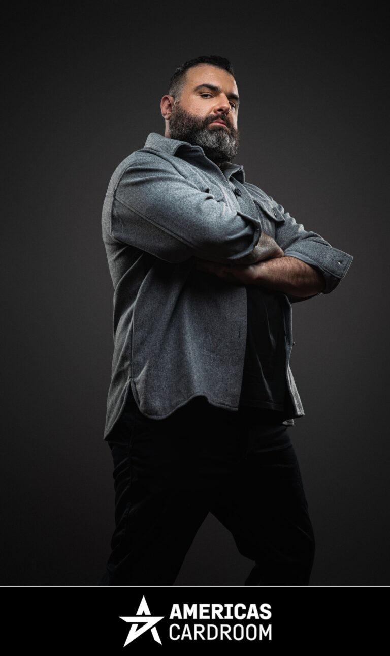 A medium shot of Jon Van Fleet, an ACR poker pro, with a full beard, looking intently towards the camera with his arms crossed. He is wearing a grey collared shirt over a dark t-shirt and black pants, against a dark background.