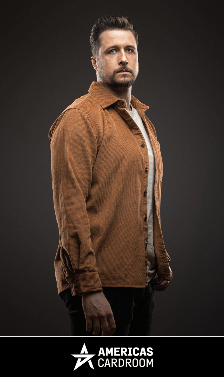 A medium shot of Jon Pardy, an ACR poker pro, with a beard, looking intently forward. He is wearing an open brown corduroy collared shirt over a light-colored t-shirt and dark pants, against a dark background.
