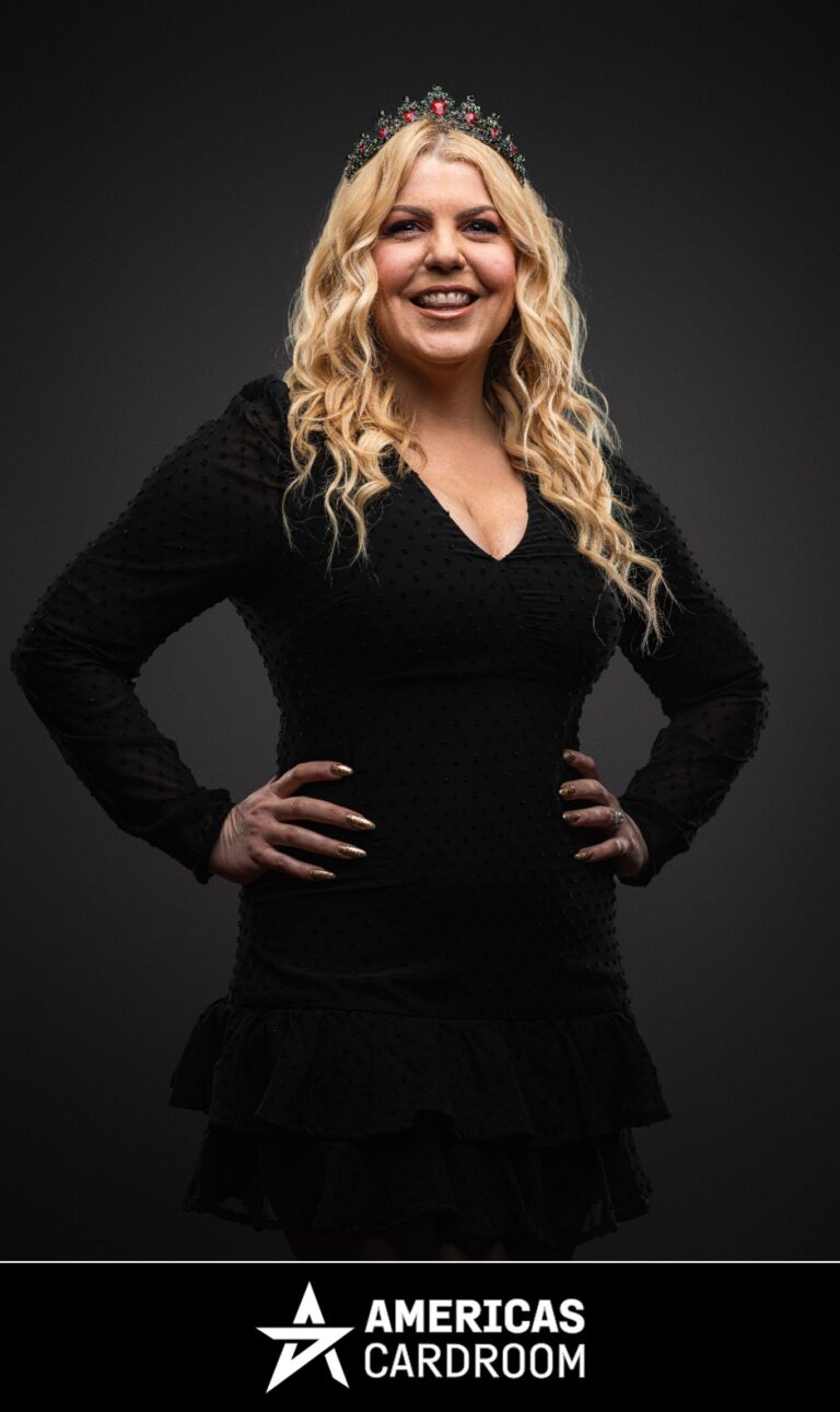 A medium shot of Katie Lindsay, an ACR poker pro, smiling widely with her hands on her hips. She has long, wavy blonde hair and is wearing a festive red and green jeweled crown, a black dress with sheer long sleeves and textured dots, and a ring on her left hand, against a dark background. Her nails are painted gold.