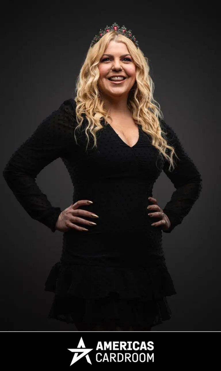 A medium shot of Katie Lindsay, an ACR poker pro, smiling widely with her hands on her hips. She has long, wavy blonde hair and is wearing a festive red and green jeweled crown, a black dress with sheer long sleeves and textured dots, and a ring on her left hand, against a dark background. Her nails are painted gold.