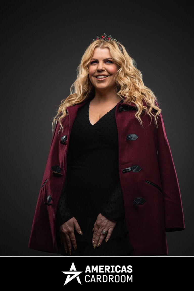 A medium shot of Katie Lindsay, an ACR poker pro, smiling and looking slightly to her left. She has long, wavy blonde hair and is wearing a festive red and gold headpiece, a dark red coat with toggle closures, over a black dress, against a dark background. Her nails are painted gold.