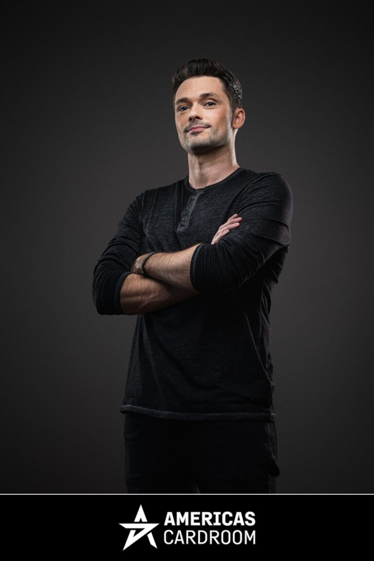 A full-body shot of Michael Loncar, an ACR poker pro, standing with his arms crossed and a slight smile. He is wearing a long-sleeved dark grey Henley shirt and dark pants, against a dark background.