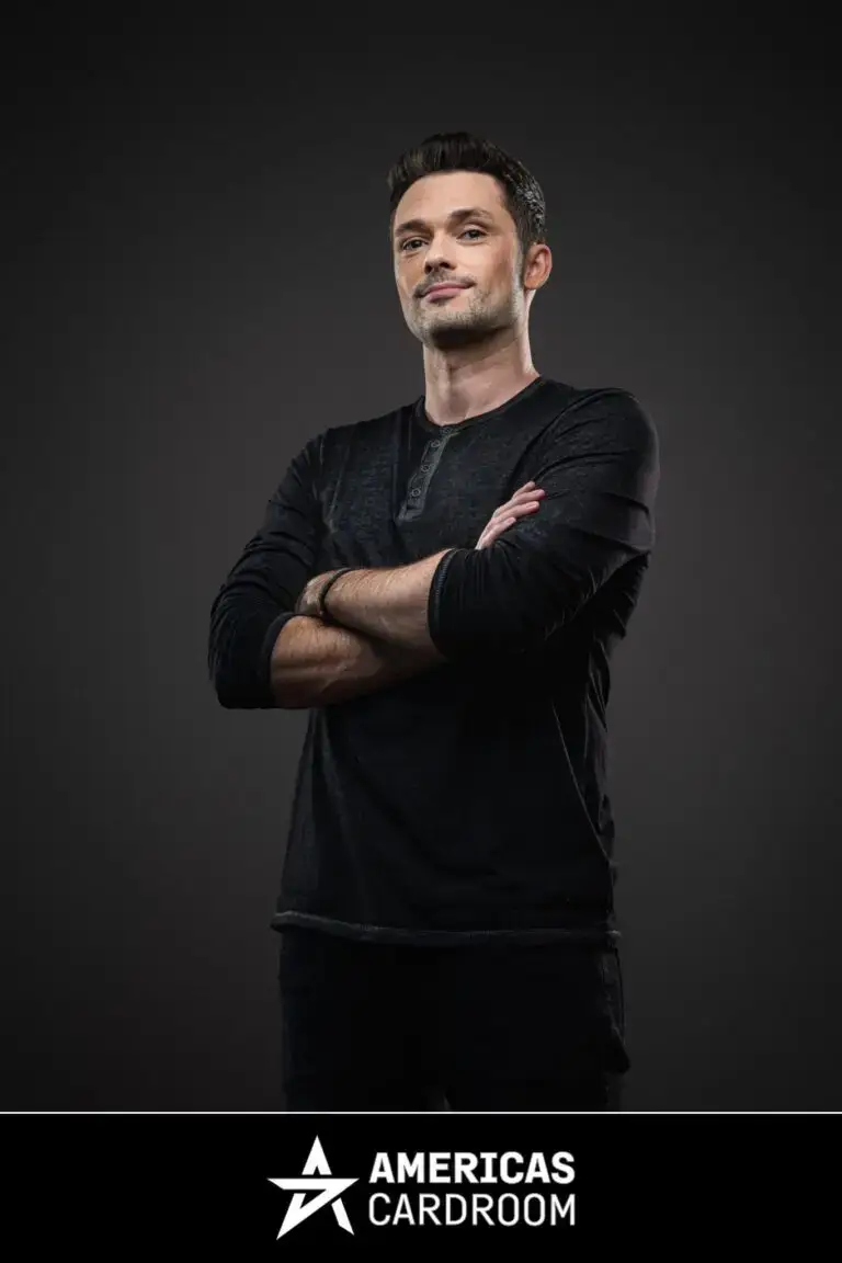 A full-body shot of Michael Loncar, an ACR poker pro, standing with his arms crossed and a slight smile. He is wearing a long-sleeved dark grey Henley shirt and dark pants, against a dark background.