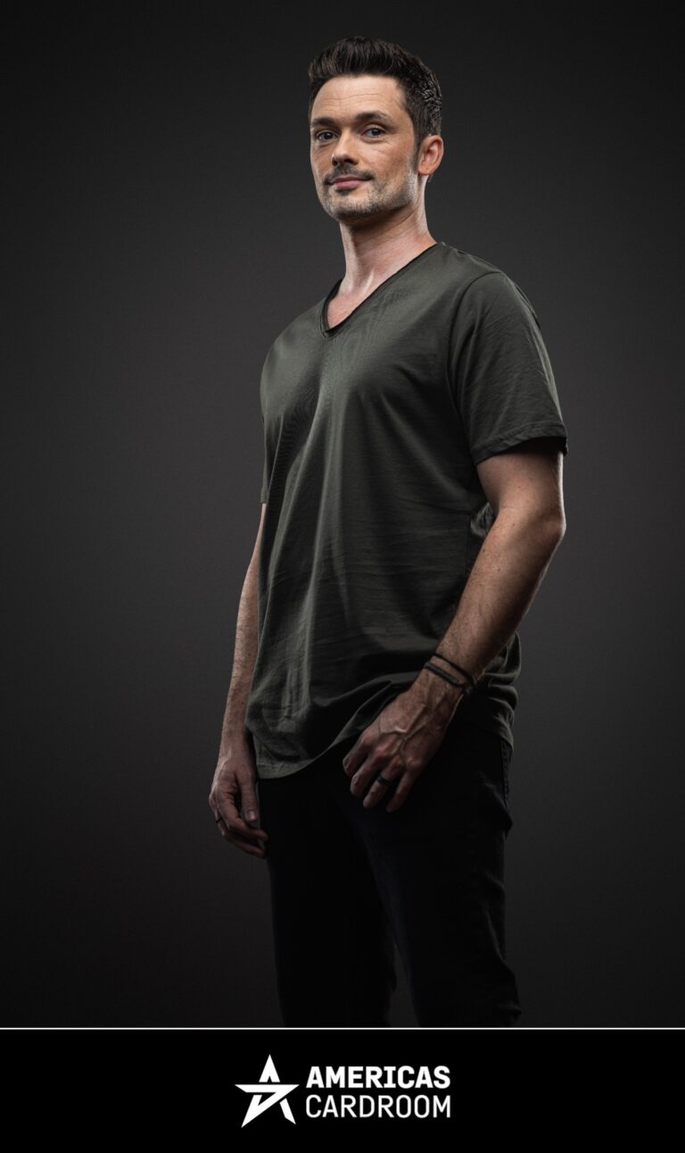 A three-quarter shot of Michael Loncar, an ACR poker pro, looking directly at the camera with a serious expression. He is wearing a dark olive green V-neck t-shirt, dark pants, and a black bracelet on his left wrist, against a dark background.