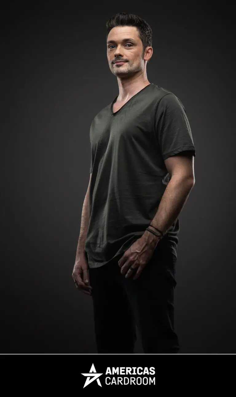 A three-quarter shot of Michael Loncar, an ACR poker pro, looking directly at the camera with a serious expression. He is wearing a dark olive green V-neck t-shirt, dark pants, and a black bracelet on his left wrist, against a dark background.