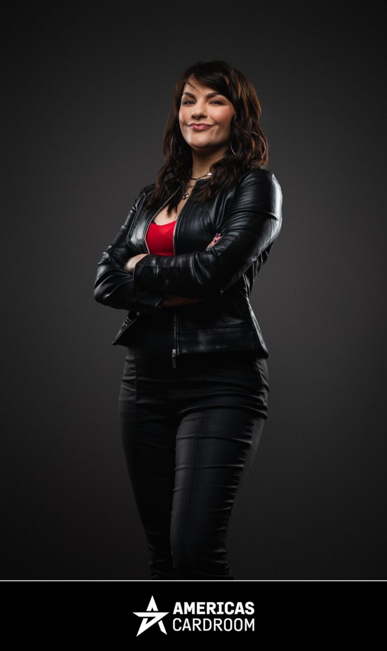 A medium shot of Monika Zukowicz, an ACR poker pro, standing with her arms crossed and a slight smile. She has dark, wavy hair and is wearing a black leather jacket over a red top and black pants, against a dark background.