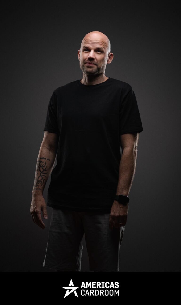 A medium shot of Owen Gaines, an ACR poker pro, with a shaved head and a light beard, looking intently towards the camera. He is wearing a plain black t-shirt, dark shorts, and a black watch on his left wrist, with tattoos visible on his left arm, against a dark background.