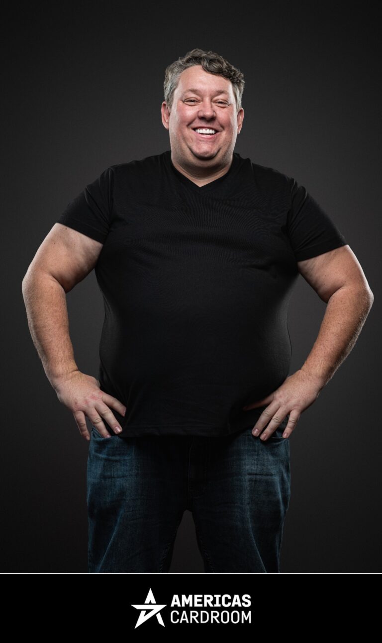 A medium shot of Rene Nezhoda, an ACR poker pro, standing with his hands on his hips and a wide, joyful smile. He is wearing a plain black t-shirt and blue jeans, against a dark background.