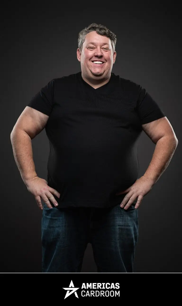 A medium shot of Rene Nezhoda, an ACR poker pro, standing with his hands on his hips and a wide, joyful smile. He is wearing a plain black t-shirt and blue jeans, against a dark background.