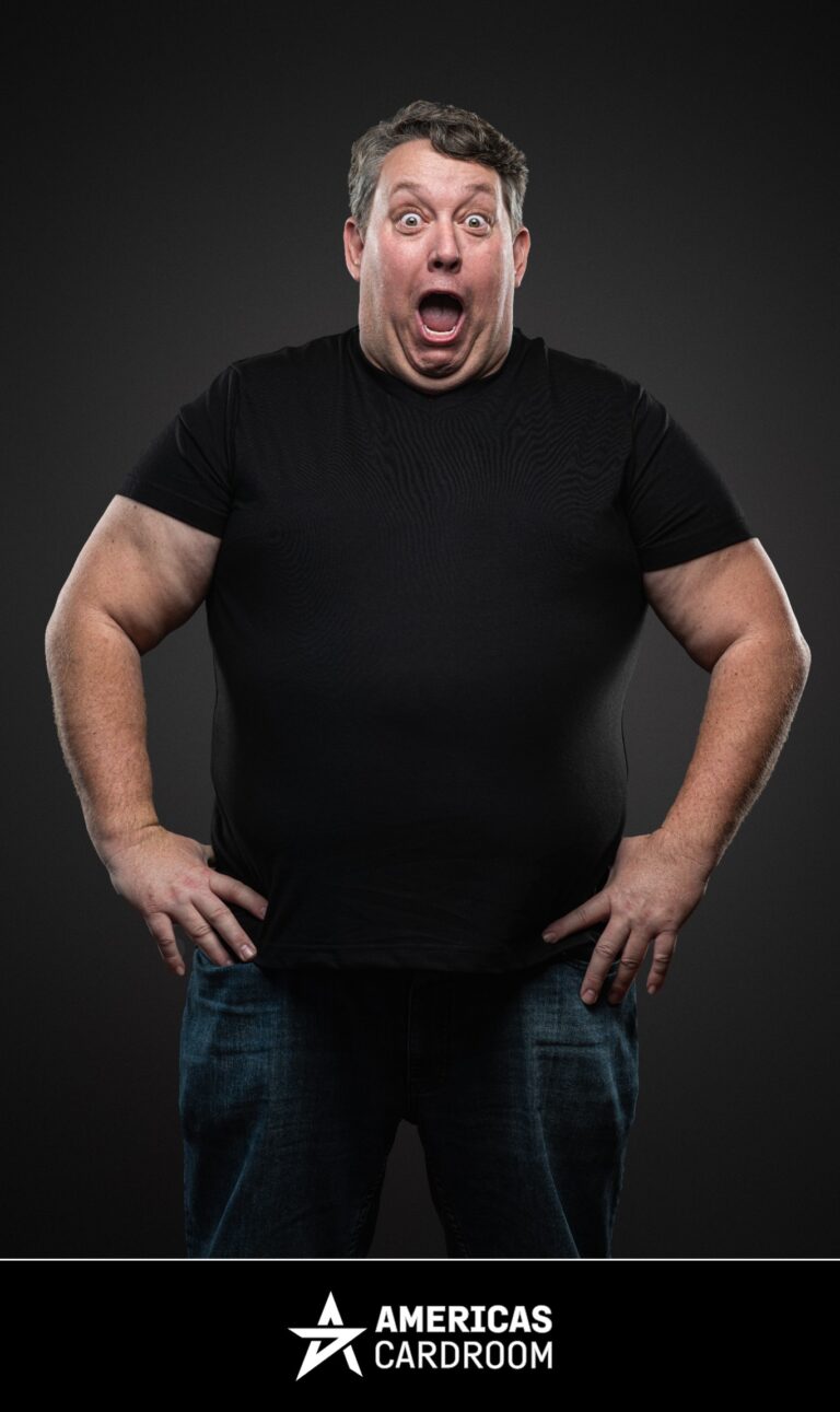 A medium shot of Rene Nezhoda, an ACR poker pro, with a look of exaggerated surprise on his face, mouth agape and eyes wide. He is standing with his hands on his hips, wearing a plain black t-shirt and blue jeans, against a dark background.