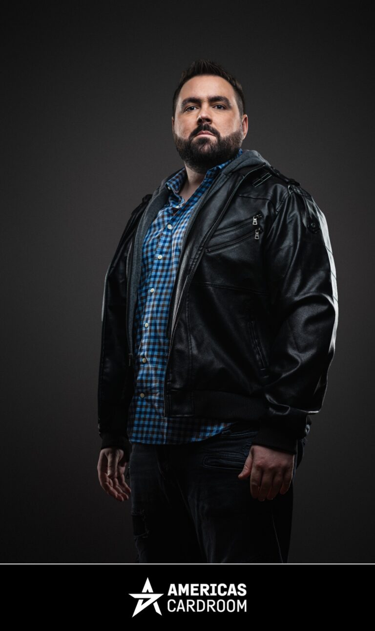 A medium shot of Robert Kuhn, an ACR poker pro, looking directly at the camera with a serious expression. He has a beard and is wearing a black leather jacket over a blue and white plaid shirt, against a dark background.