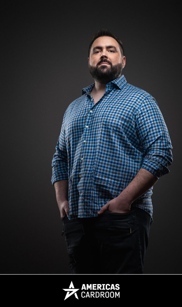A medium shot of Robert Kuhn, an ACR poker pro, standing with his hands in his pockets, looking forward with a neutral expression. He is wearing a blue and brown plaid button-up shirt and dark jeans, against a dark background.