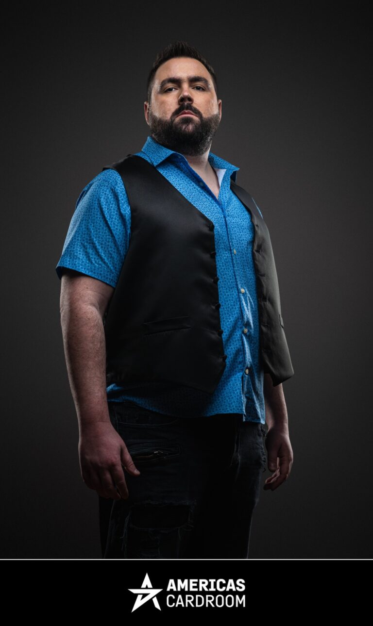 A medium shot of Robert Kuhn, an ACR poker pro, standing with a serious expression, wearing a black vest over a bright blue short-sleeved button-up shirt, against a dark background.