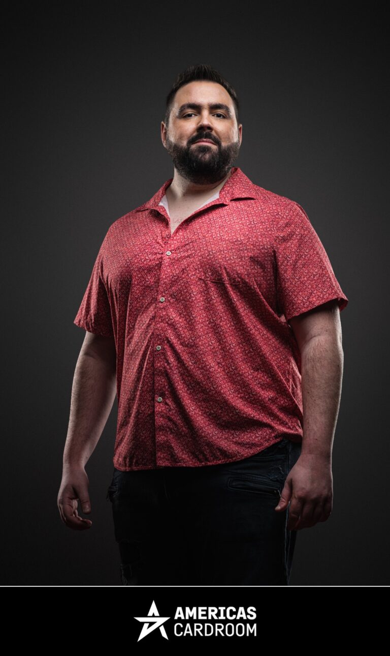 A medium shot of Robert Kuhn, an ACR poker pro, standing with a neutral expression, wearing a red short-sleeved button-up shirt with a subtle pattern, against a dark background.