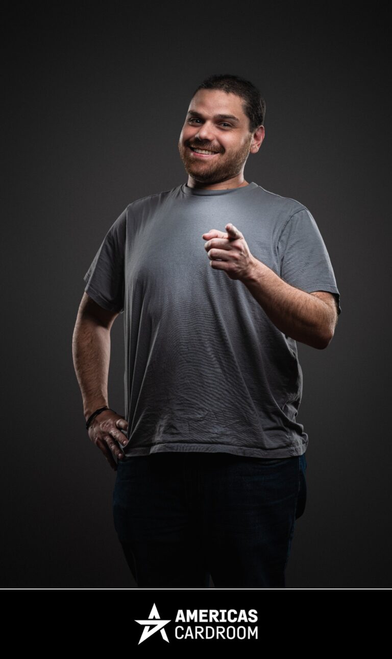 A medium shot of Robert Kuhn, an ACR poker pro, smiling and pointing forward with his right hand, while his left hand is on his hip. He is wearing a plain grey t-shirt and dark pants, against a dark background.