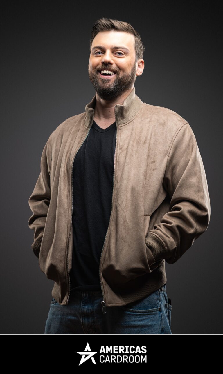 A medium shot of Ryan Depaulo, an ACR poker pro, with a beard, smiling and looking up, wearing a light brown or tan bomber jacket over a black V-neck shirt and blue jeans, with his hands in his pockets, against a dark background.