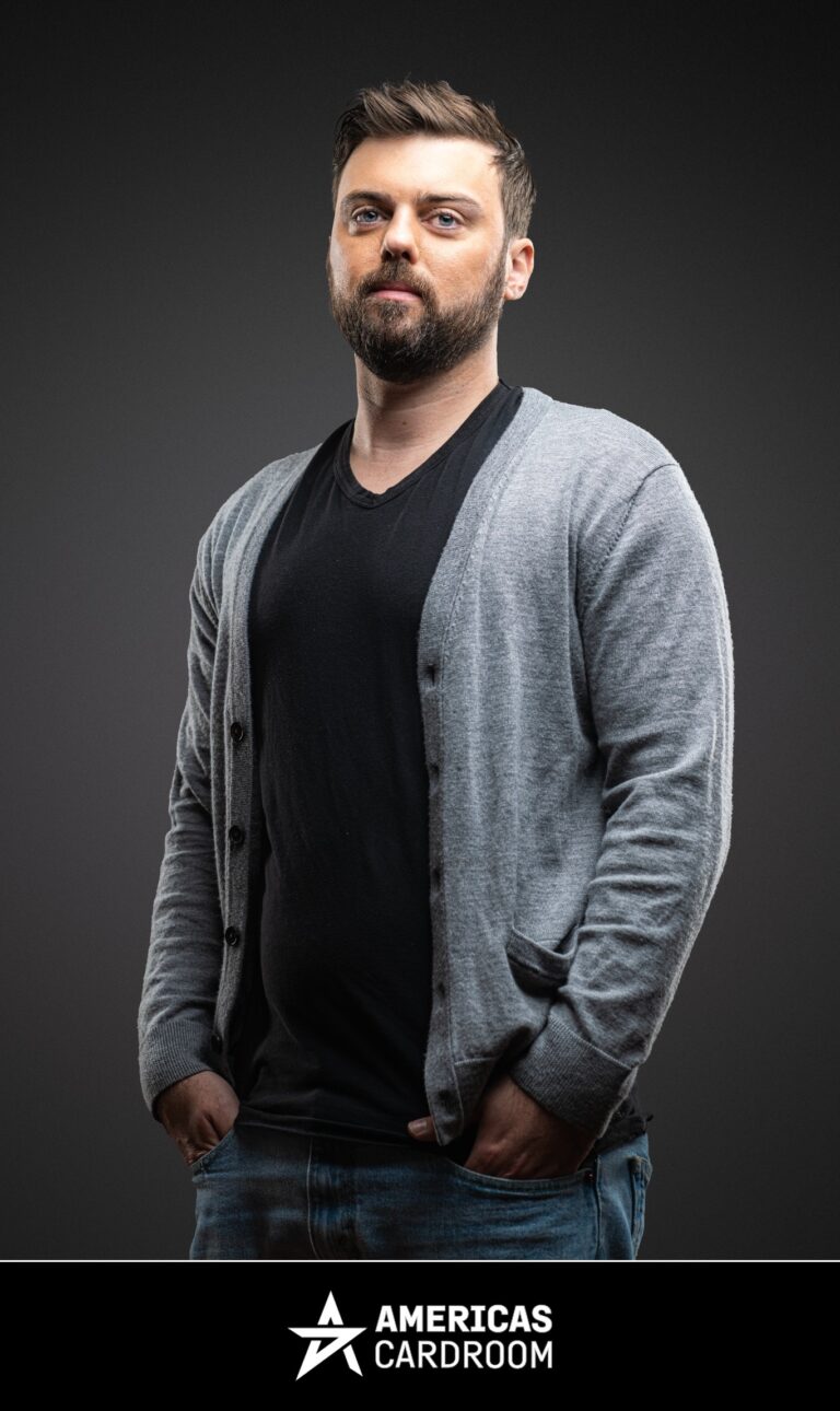 A medium shot of Ryan Depaulo, an ACR poker pro, with a beard, looking seriously at the camera, wearing a light grey button-up cardigan over a black V-neck t-shirt and blue jeans, with his hands in his pockets, against a dark background.