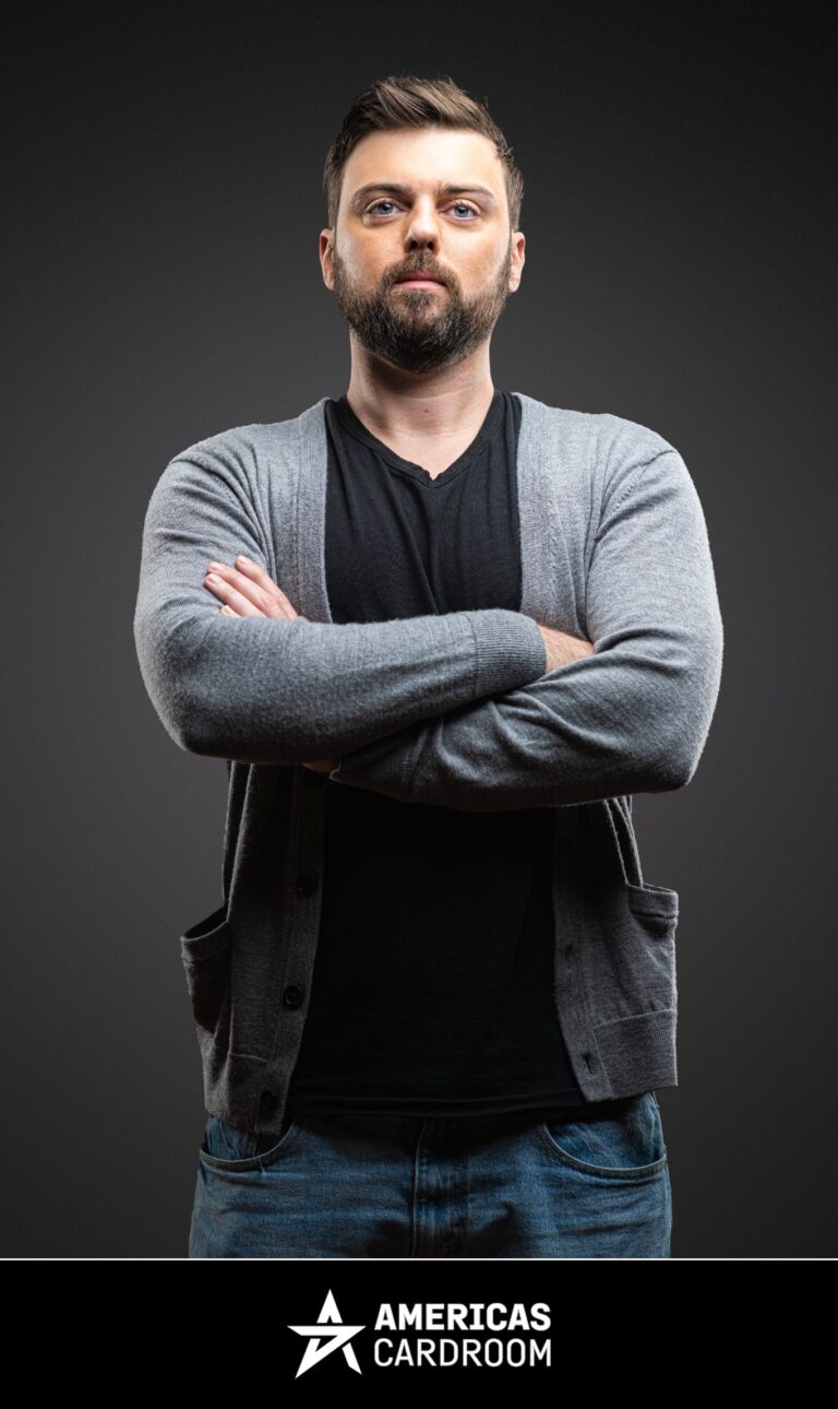A medium shot of Ryan Depaulo, an ACR poker pro, with a beard, looking intensely at the camera with his arms crossed, wearing a light grey button-up cardigan over a black V-neck t-shirt and blue jeans, against a dark background.