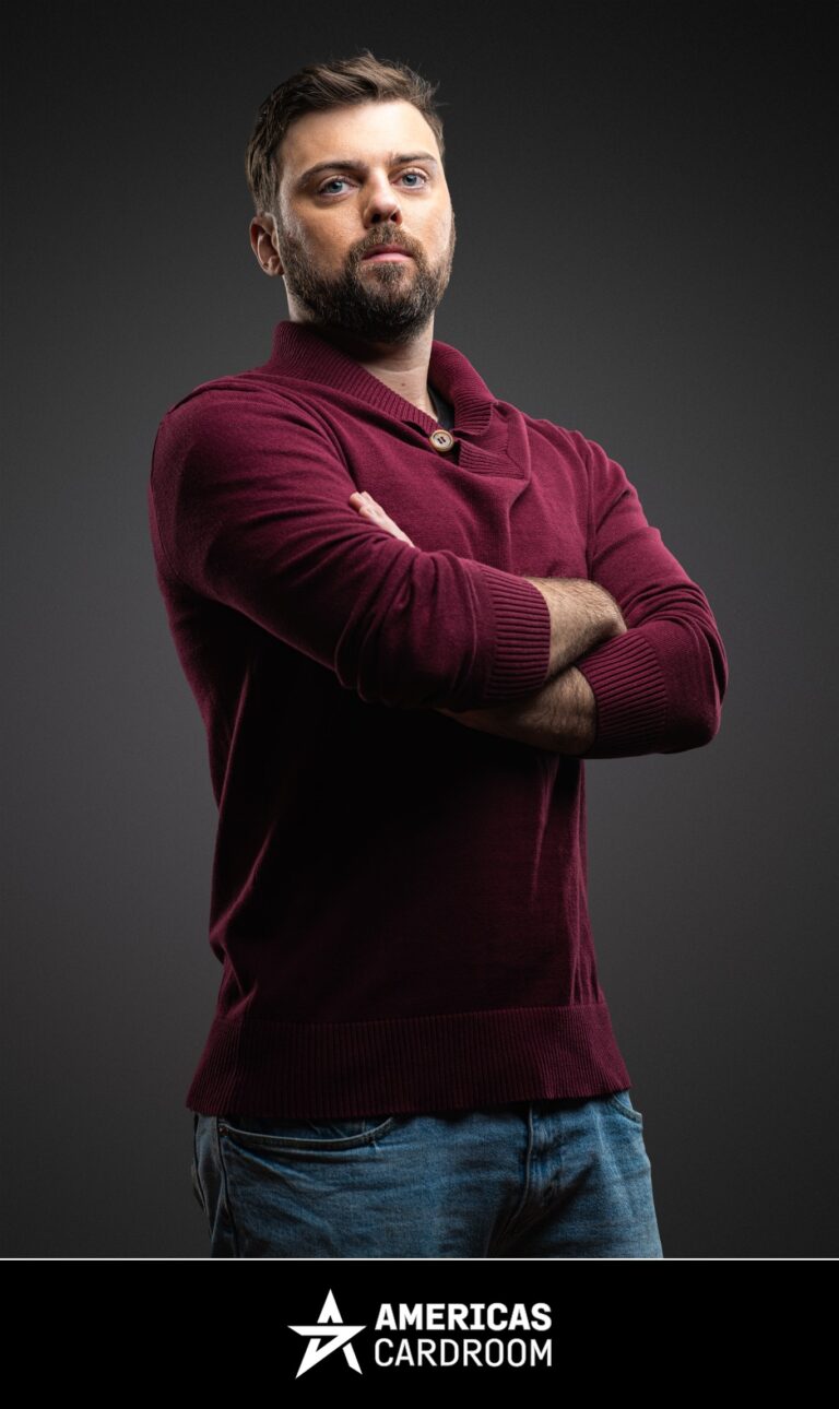 A medium shot of Ryan Depaulo, an ACR poker pro, with a beard, looking at the camera with his arms crossed, wearing a long-sleeved maroon collared sweater with a single button, and blue jeans, against a dark background.